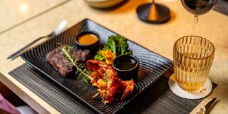 A gourmet dish featuring lobster, steak, and colorful vegetables, complemented by sauces in small bowls and a stylish glass of drink.