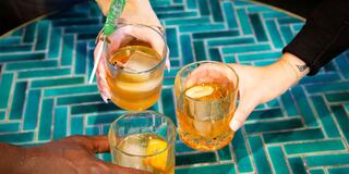 Four hands hold drinks on a vibrant teal table, creating a festive atmosphere with glasses garnished with citrus.