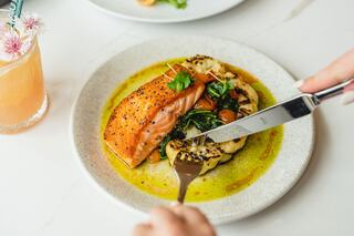 A beautifully plated salmon fillet atop sautéed greens and roasted cauliflower, drizzled with vibrant sauce, accompanied by a refreshing drink.
