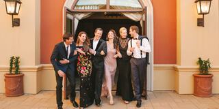 A group of six elegantly dressed people joyfully walking together outside a building with stylish decor and warm lighting.