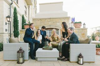 A stylish group of six people in formal attire enjoy drinks and laughter on an outdoor seating area, surrounded by elegant decor.