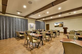 A spacious meeting room with tables and chairs, wooden beams on the ceiling, and large curtains. A podium and screen are set up for presentations.