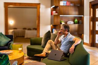 A man sits comfortably on a green sofa, speaking on the phone while working on his laptop in a stylish, modern lounge setting.