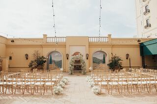 A charming outdoor wedding venue with golden chairs arranged in rows, adorned with floral decor, and a backdrop featuring arched structures.