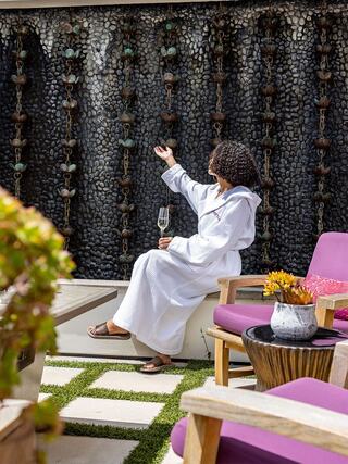 A woman in a cozy robe enjoys a drink while relaxing by a stone wall adorned with water features, surrounded by stylish seating.