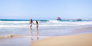 Two girls wade in the shallow water, holding hands, with a picturesque pier and clear blue sky in the background on a sunny beach day.