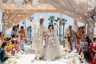 A joyful couple celebrates their wedding under a floral arch, surrounded by family and friends, with the beach in the background.