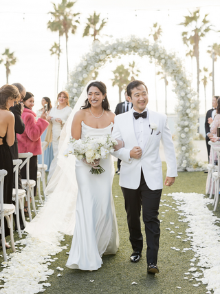 A couple in formal attire stands together, with a stylish dog in a bowtie sitting in front, adding charm to the scene.