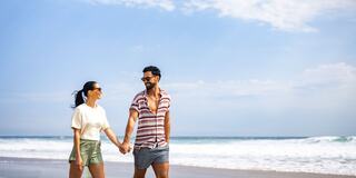 A couple strolls hand-in-hand along a beach, smiling at each other, with the ocean and a clear sky in the background.