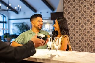A couple smiles at each other while a server presents a bottle of champagne beside elegant glasses on a marble counter.