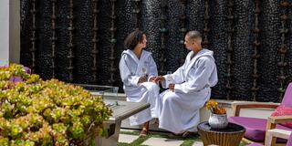 Two people in cozy robes sit together in a tranquil outdoor setting, enjoying drinks and engaging in a warm conversation amidst greenery.