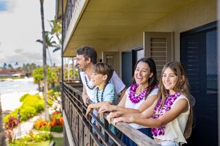 A cheerful family of four, wearing leis, enjoys a scenic view from their balcony, smiling and sharing a joyful moment together.