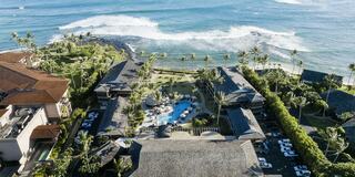 A coastal resort features palm trees, a pool, and ocean waves crashing nearby, creating a tranquil tropical atmosphere.