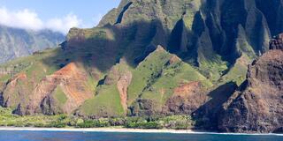 Lush green mountains rise dramatically from a coastal shoreline, featuring steep cliffs and vibrant landscapes under a clear blue sky.