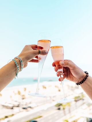 Two hands raise sparkling glasses filled with pink champagne against a vibrant sky, celebrating with a beautiful beach view in the background.
