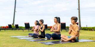 Four individuals practice yoga on mats in a serene outdoor setting, wearing headphones and focusing in a peaceful environment.