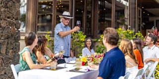 A group of friends gathers around a table, enjoying drinks and laughter, as a man raises a glass for a toast amidst a garden setting.