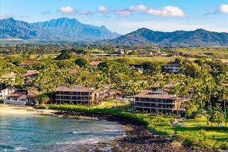 Tropical beachfront resort with palm trees, lush greenery, and majestic mountains in the background under a clear blue sky.