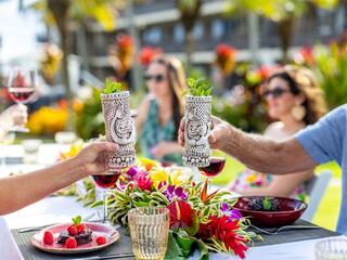 Two people toast with decorative glasses amidst a vibrant outdoor setting, featuring tropical flowers and a cheerful gathering of friends.