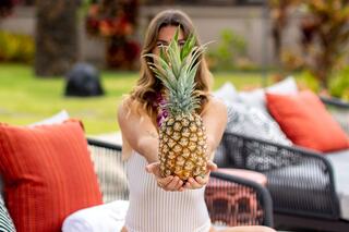 A person in a swimsuit holds a pineapple in front of them, surrounded by colorful cushions in a lush outdoor setting.