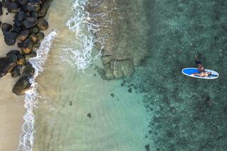 Aerial view of a clear turquoise sea with a surfer on a paddleboard, rocky shore, and gentle waves lapping against the beach.