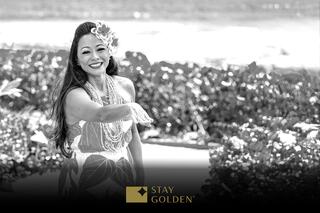 A woman in a colorful dress adorned with flowers smiles and gestures joyfully against a scenic backdrop, embodying a warm, inviting spirit.