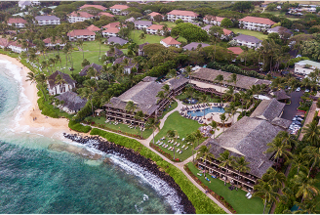 Aerial view of a tropical resort near the beach, featuring lush greenery, sandy shores, and homes surrounded by palm trees.