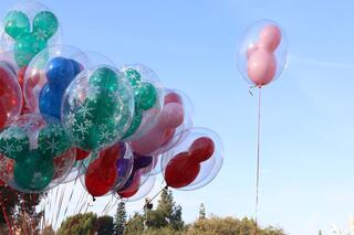 A cluster of colorful, Mickey Mouse-shaped balloons floats against a clear blue sky, featuring vibrant red, green, and pink hues.