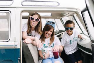 Three kids playfully pose in a vintage van, wearing colorful sunglasses and matching shirts, making silly faces and enjoying drinks.