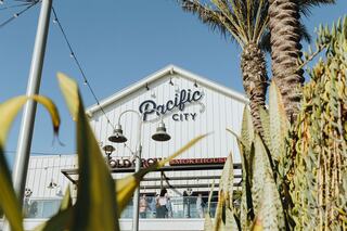 A bustling coastal venue named Pacific City features palm trees and a smokehouse, inviting guests to enjoy the vibrant atmosphere.