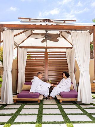 A couple in cozy robes relax in a romantic cabana, enjoying each other's company amidst lush greenery and a serene atmosphere.