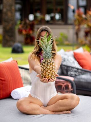 A woman in a striped swimsuit holds a pineapple out in front of her while sitting cross-legged on a lounge chair amid colorful cushions.