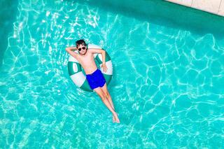 A boy relaxes on a buoy in a shimmering pool, wearing sunglasses and blue swim trunks, enjoying a sunny day.