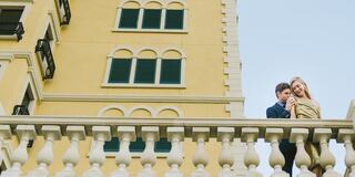 A couple stands on a balcony, enjoying a moment together against a backdrop of an elegant yellow building and blue sky.