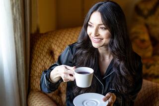 A woman in a black robe sits comfortably in a chair, smiling while holding a cup of tea, enjoying a serene moment by the window.