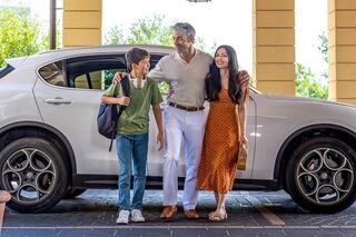 A family stands happily by a car, with a boy in casual clothes and a man and woman dressed elegantly, enjoying a cheerful moment together.