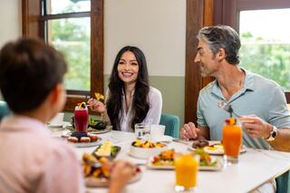 A family enjoys a lively meal together, sharing smiles and dishes in a bright, inviting space with lush greenery outside.