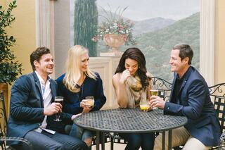 Four friends enjoy drinks at a stylish outdoor table, laughing and conversing against a picturesque backdrop of nature and architecture.
