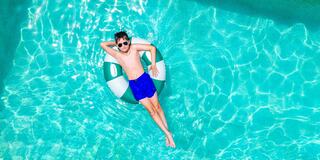 A boy lounges on a pool float, wearing sunglasses and blue shorts, enjoying a sunny day in sparkling turquoise water.