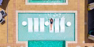 A woman relaxes on floating loungers in a bright blue pool, surrounded by sunlit tiles and lounge chairs. It's a serene summer scene.