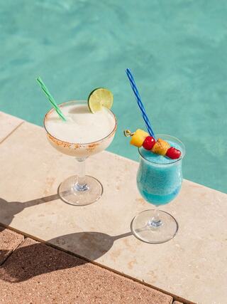 Two colorful cocktails sit by a shimmering pool, one a creamy margarita with a lime, the other a vibrant blue drink topped with fruit.