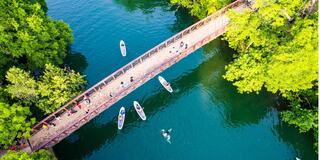 Aerial view of a bridge over a vibrant blue river, with people walking and kayaking amidst lush greenery.