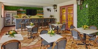 A cozy café features tables with flowers, rattan chairs, and a bar stocked with drinks, set against a backdrop of vibrant green foliage.