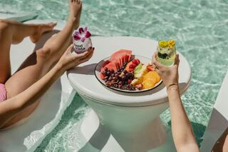 Two hands hold colorful drinks beside a circular table with a vibrant fruit platter, set against a sparkling pool backdrop.