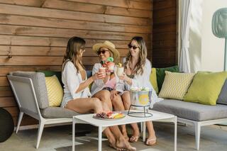 Three friends in summer attire sit on a cozy outdoor couch, enjoying colorful drinks and sharing laughter in a relaxed atmosphere.