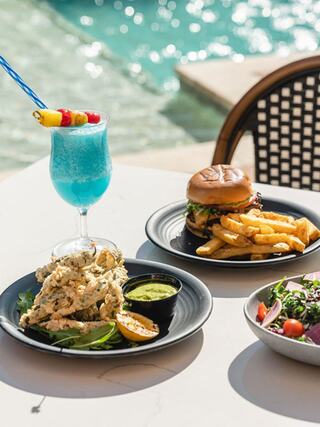 A vibrant meal featuring a burger with fries, salad, and a colorful cocktail, set against a sunny poolside backdrop.
