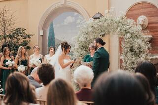 A couple exchanges vows in a romantic setting, surrounded by guests and adorned with a floral arch, creating a heartfelt wedding scene.