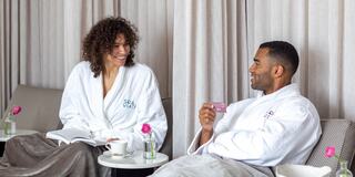 Two smiling individuals in spa robes relax together, enjoying drinks and conversation in a cozy, serene setting.