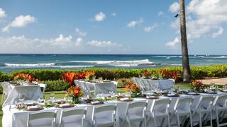 A picturesque outdoor setting features elegantly set tables on lush green grass, with vibrant plants and a stunning ocean backdrop.