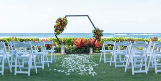 A serene outdoor wedding setup features white chairs, floral decorations, and a scenic ocean backdrop, creating a romantic atmosphere.